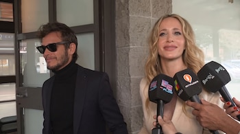 Una mujer de cabello claro frente a tres micrófonos junto a un hombre con gafas oscuras y traje frente a una pared.