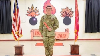 Hombre joven de pie con uniforme militar de camuflaje, sosteniendo un cilindro naranja. Dos banderas y emblemas de bombas con llamas decoran el fondo