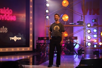 Un hombre sonríe en un escenario con focos de iluminación, sillas de director y pantallas con luces de neón y logotipos