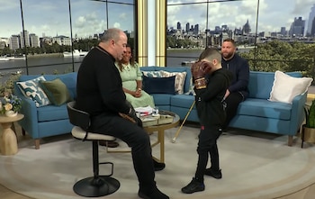 Un hombre en una silla alta observa a un niño con guantes de boxeo. Una mujer y otro hombre están en un sofá. Una mesa redonda se ve. Ventanales con vista a la ciudad