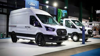 Dos furgonetas utilitarias blancas Ford Transit exhibidas en una exposición, con el logo Ford Pro y un soporte azul con un código QR