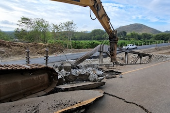 Oruga y brazo de una excavadora sobre escombros de hormigón y asfalto agrietado cerca de una carretera con un vehículo blanco al fondo.