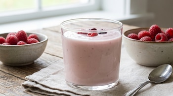 Un vaso de yogur rosa claro con algunas bayas en la superficie, flanqueado por dos cuencos con frambuesas, una cuchara y un paño de lino sobre una mesa de madera.