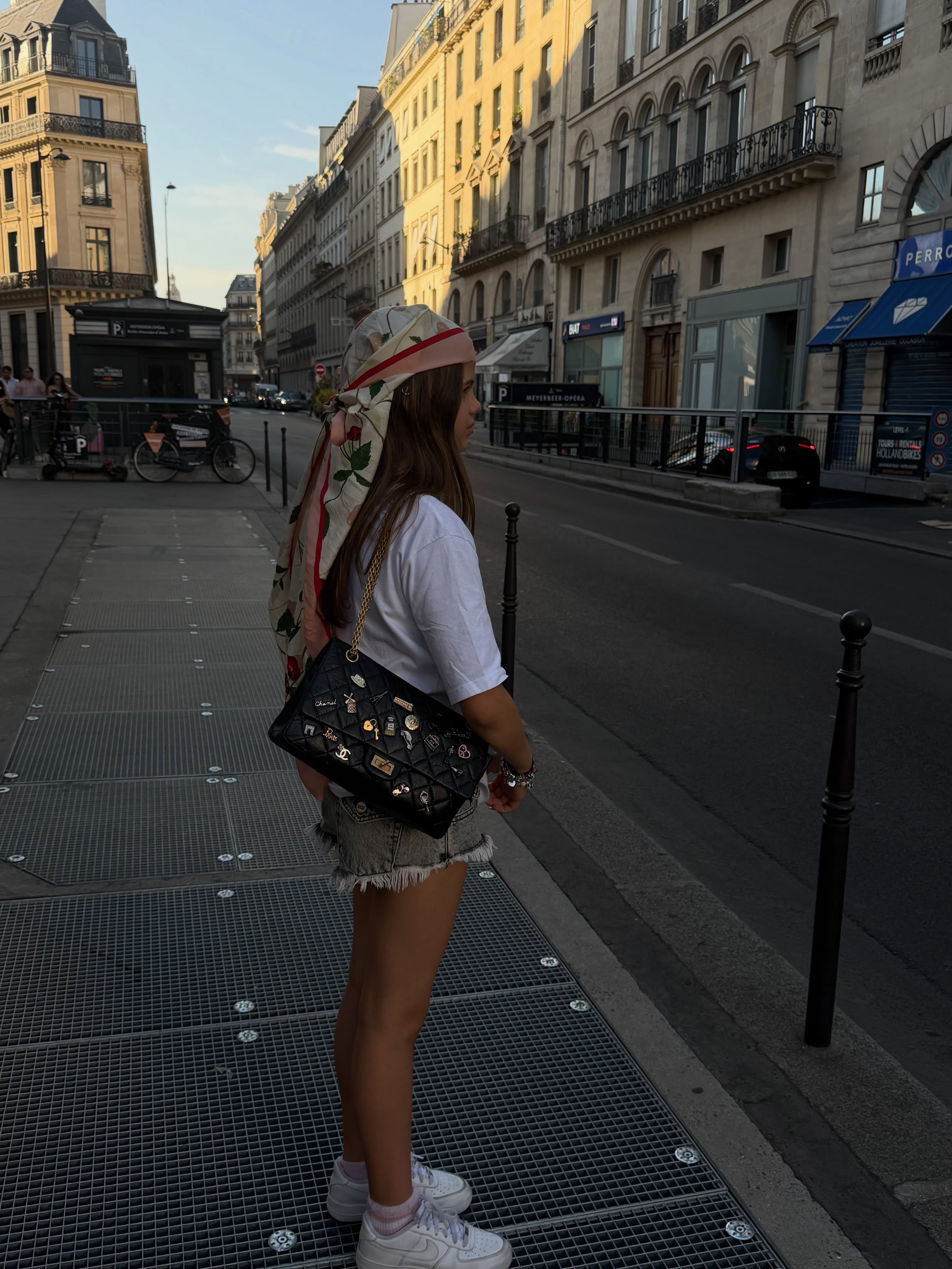 Francesca lució un pañuelo estampado y cartera decorada camina por una calle de París, con edificios históricos y vehículos estacionados de fondo.