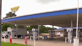 Vista exterior de una gasolinera con dosel azul y blanco, varios surtidores, un camión blanco y un coche rojo. Un letrero de 'Waffle House' es visible al fondo