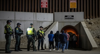 Agentes de CBP con uniformes y chalecos reflectantes escoltan a personas fuera de un túnel de drenaje de concreto en la frontera California-México por la noche.