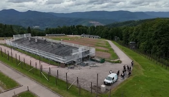 Vista aérea de un campamento rodeado por cercas y montañas; un edificio con andamios, un camino, una camioneta blanca y personas se observan en primer plano