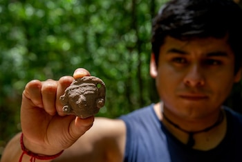 Mano de persona sostiene una pieza de cerámica marrón con forma de cabeza. Al fondo, rostro borroso de joven y vegetación verde