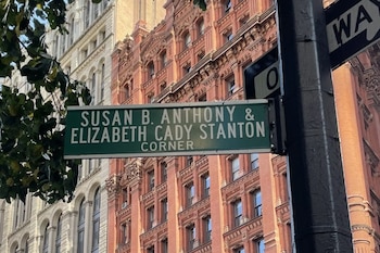Un cartel vial verde con texto blanco indica 'Susan B. Anthony & Elizabeth Cady Stanton Corner' sobre un fondo de edificios y follaje