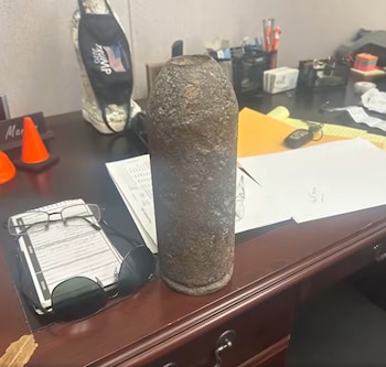 Un proyectil de artillería oxidado y corroído se erige verticalmente sobre un escritorio de madera. Hay papeles, gafas, conos y una mochila de fondo