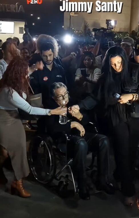 El cantante Jimmy Santi es asistido en silla de ruedas por dos mujeres en medio de una multitud de prensa, luego de un incidente al intentar ingresar al velorio de Manolo Rojas. (TikTok)