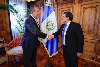 Dos hombres con traje formal se estrechan la mano ante la bandera de Guatemala en una habitación con paneles de madera