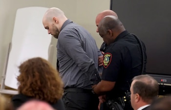 Hombre calvo y barbudo, con esposas y camisa gris, es escoltado por dos alguaciles uniformados del Condado de Montgomery, Texas, en una sala de tribunal