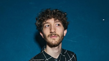 Primer plano de Teo López Puccio con cabello rizado y barba, vistiendo una camisa a cuadros blanca y negra, con un fondo azul oscuro