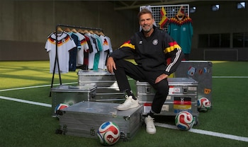Jurgen Klopp sentado en un baúl metálico en un campo de fútbol con camisetas de la selección alemana colgadas, baúles y balones