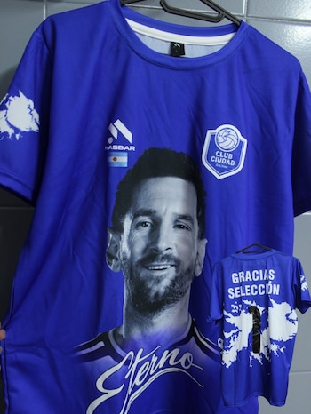 Camiseta azul con rostro de Lionel Messi, escudo Club Ciudad Bolívar, bandera argentina y texto 'Eterno'. Detrás, camiseta con 'Gracias Selección' y mapa de Argentina