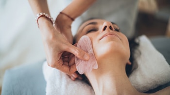Una mujer con ojos cerrados yace mientras una mano aplica una herramienta gua sha de cuarzo rosa en su mejilla. Hay una toalla blanca.