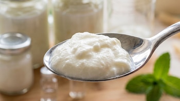 Cuchara de metal con nata coagulada blanca y cremosa. Fondo desenfocado con frascos de vidrio y una hoja de menta sobre una superficie de madera