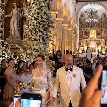 Una novia con vestido blanco y un novio de esmoquin caminan tomados de la mano en una iglesia rodeados de flores e invitados.