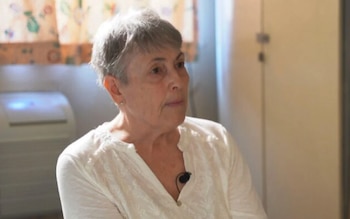 Una mujer de pelo gris corto y camisa blanca sentada en interiores, mirando hacia la derecha. Se ve un micrófono de solapa negro en su camisa