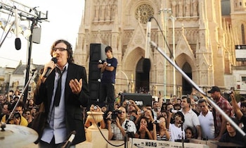 Charly García canta con un micrófono en la mano frente a una multitud y a la fachada de la Basílica de Luján