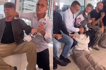 Un hombre con gafas sujeta por el rostro a dos personas que sostienen pancartas durante un evento en un espacio interior