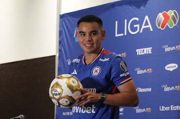 Hombre joven con camiseta azul de Cruz Azul, sonriendo y sosteniendo un balón Voit dorado y blanco, frente a un fondo azul con logos de Liga BBVA MX y patrocinadores