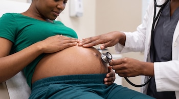Una mujer embarazada con el vientre descubierto y las manos de una profesional de la salud colocando un estetoscopio sobre su abdomen.