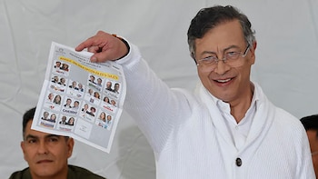 Un hombre con gafas y camisa blanca sonríe a la cámara mientras sostiene una boleta electoral blanca con múltiples fotografías de candidatos