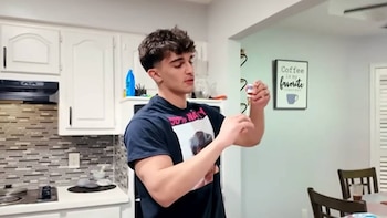Joven de cabello oscuro y camiseta negra sostiene un pequeño vial blanco y rojo. Se ven gabinetes blancos y un salpicadero de azulejos en el fondo