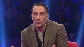 Primer plano de Luciano Cáceres, un hombre con ojos azules y cabello oscuro rizado, vistiendo una chaqueta de tweed marrón y camisa clara, hablando en un micrófono. Fondo con luces azules