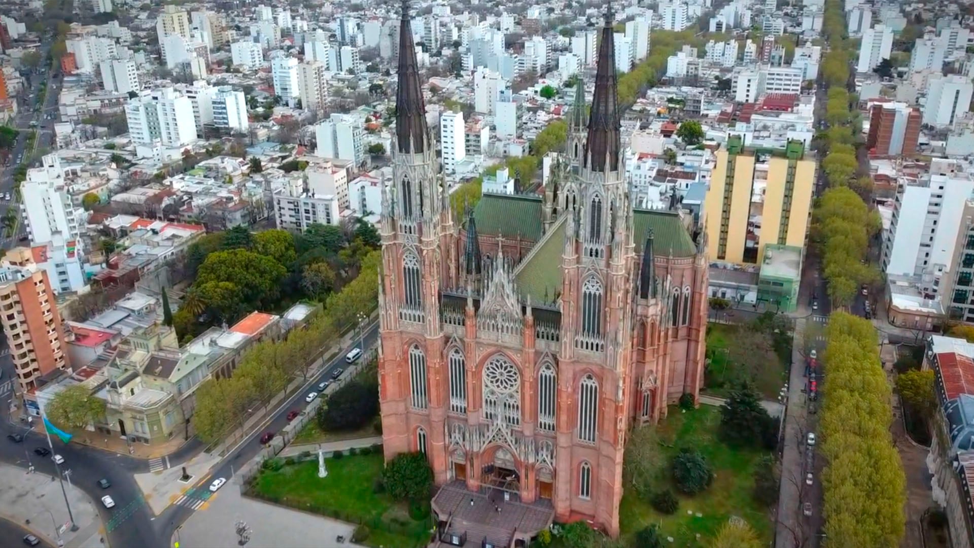 Desde una perspectiva elevada, la imponente Catedral de La Plata se alza en el paisaje urbano, destacando su arquitectura neogótica entre las calles arboladas y edificios de la ciudad argentina. (Captura de video)