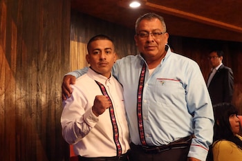 Dos hombres: uno con camisa blanca bordada y puño cerrado; otro con camisa azul bordada, lentes y brazo sobre el hombro del primero. Fondo de madera