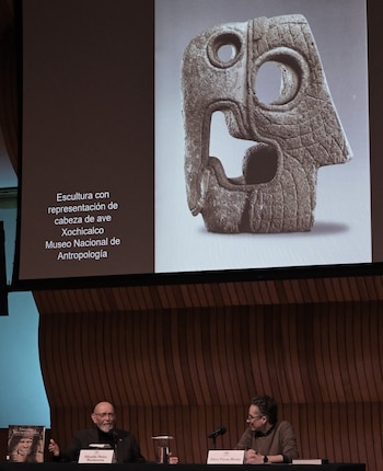Dos hombres sentados en un escenario frente a una pantalla con una escultura de piedra prehispánica y texto, con una pared de madera detrás