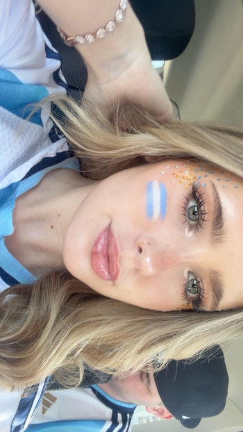 Mujer rubia con camiseta de Argentina, maquillaje facial azul y blanco con brillos, ojos maquillados, labios rosas y un brazalete