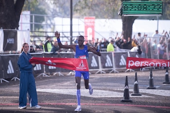 Un corredor con vestimenta azul cruza una cinta roja de meta sostenida por una mujer durante una competencia deportiva al aire libre