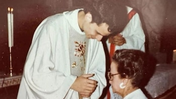 Un sacerdote joven con vestiduras blancas se inclina ante una mujer en el interior de una iglesia junto a velas encendidas