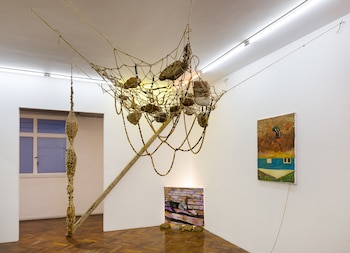 Interior de galería con paredes blancas y suelo de madera. Una red con piedras y hilos de cuentas cuelga del techo. Dos pinturas en la pared y una en el suelo