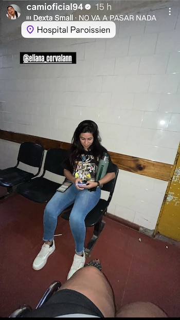 Mujer joven sentada en un banco de hospital, mirando su teléfono y sosteniendo un termo. Viste camiseta estampada y jeans azules. Pared de azulejos blancos y suelo rojo