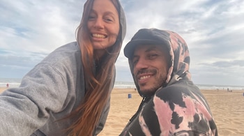Una mujer con sudadera gris y un hombre con sudadera tie-dye sonríen a la cámara; detrás se ve una playa con algunas personas y el mar bajo un cielo nublado.