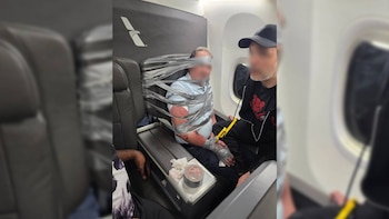 Un hombre permanece sujetado con cinta adhesiva a un asiento de avión, con un rollo de cinta sobre el apoyabrazos