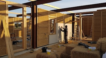 Guardería infantil en construcción con paredes de paja, estructura de madera y acero. Un trabajador aplica un revoque. Andamios, herramientas y balas de paja.