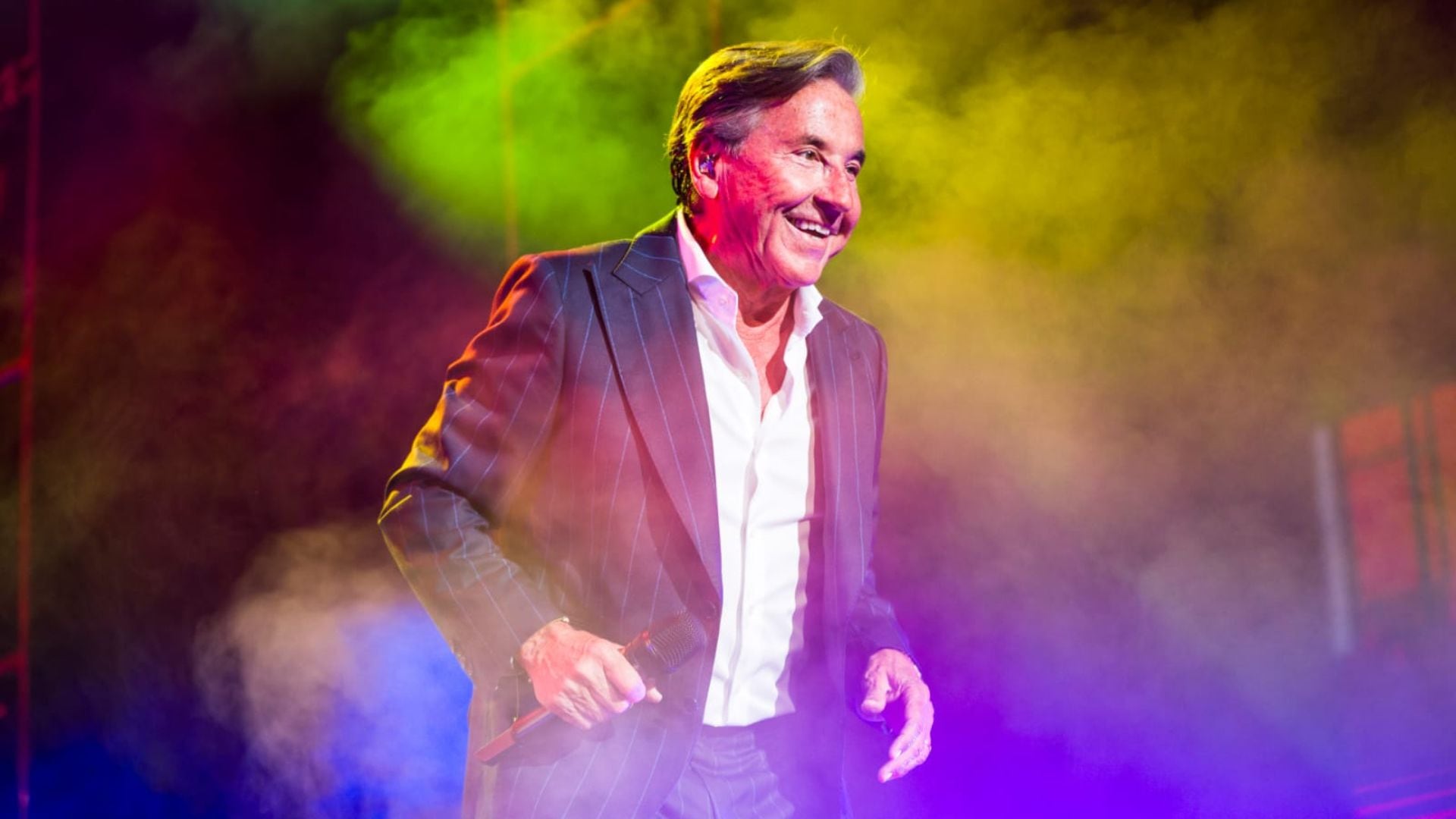 El icónico Ricardo Montaner sonríe y sostiene un micrófono en el escenario, enviando un mensaje especial a sus fans peruanos previo a su esperado concierto.