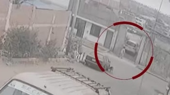 Imagen de seguridad granulada de un auto blanco en una cochera, una persona y un vehículo grande en primer plano. Edificios a los lados. Círculo rojo resalta la escena