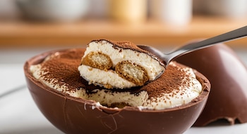 Un huevo de Pascua de chocolate cortado por la mitad, relleno de cremoso tiramisú con bizcochos, mientras una cuchara extrae una porción. Se aprecia cacao en polvo.