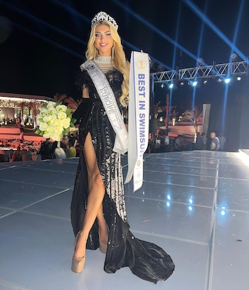 Mujer rubia con vestido negro de lentejuelas, corona, ramo de flores blancas y bandas de certamen de belleza posa en un escenario iluminado