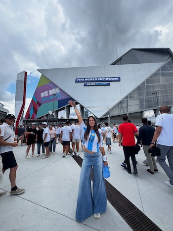 Una mujer con camiseta de Argentina y jeans acampanados levanta el brazo frente a un estadio con el letrero "FIFA WORLD CUP 2026", entre personas