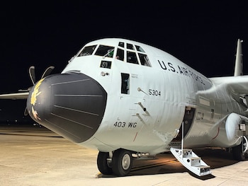 Parte frontal de un avión militar gris iluminado de noche en una pista de aterrizaje, con las escaleras de la puerta abiertas