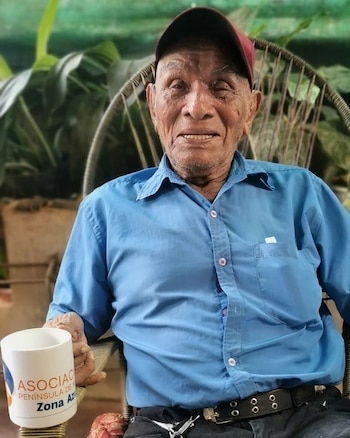 Hombre de edad avanzada sentado en una silla, con camisa azul y gorra roja. Sostiene una taza blanca con el texto "ASOCIAC PENÍNSULA DE Zona Az"