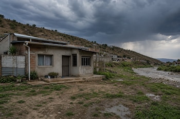 Casa de un piso construida con ladrillo y cemento junto a un camino de tierra, un río y un cerro bajo un cielo nublado.